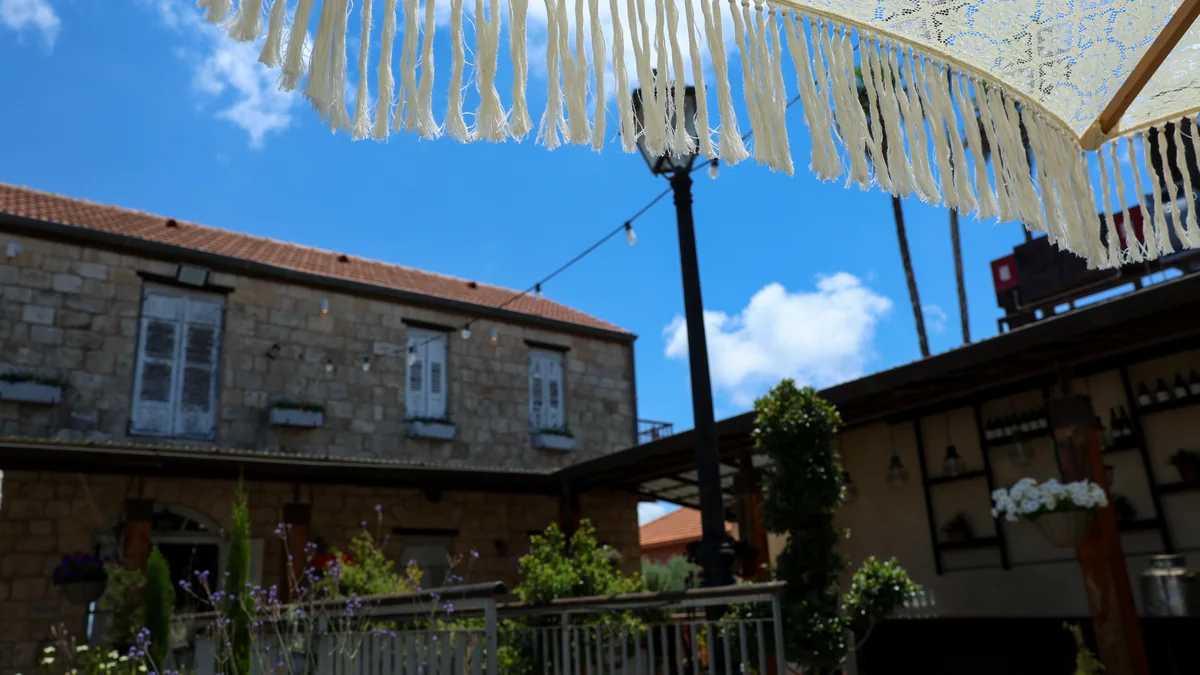 גגון בד עם פרנזים מעל רחבה חיצונית, ומבנה אבן ודקלים ברקע